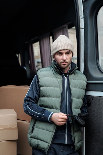 A young male mover with long, wavy hair and a black bandana is sitting in the driver’s seat of a white cargo van branded with the text 'MOVING COMPANY LOCAL & LONG DISTANCE' on the side panel. He is