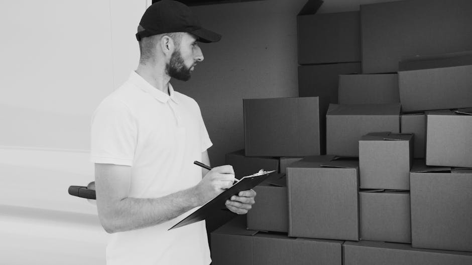 A man with a serious expression, wearing a light grey knit beanie and a padded green and navy jacket, stands at the side of a moving van during a home relocation. The van's open side door reveals a da
