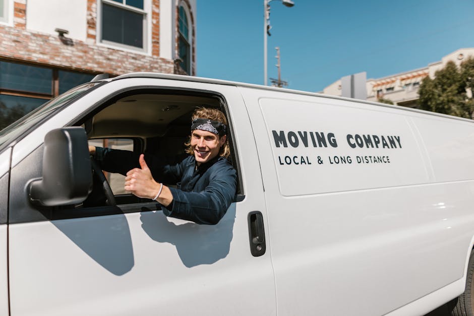 A man wearing a white t-shirt and jeans is seen outside a residential property, loading cardboard boxes into the open rear doors of a white moving van parked on the street. The boxes vary in size and 
